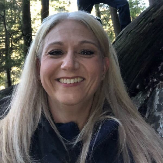 A woman smiles in front of a tree in the woods.