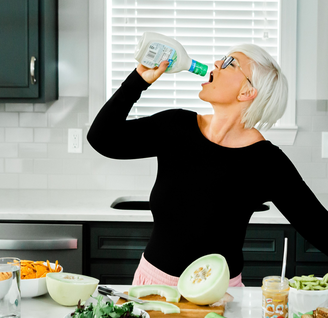 A woman drinking from a bottle in front of a kitchen full of food.