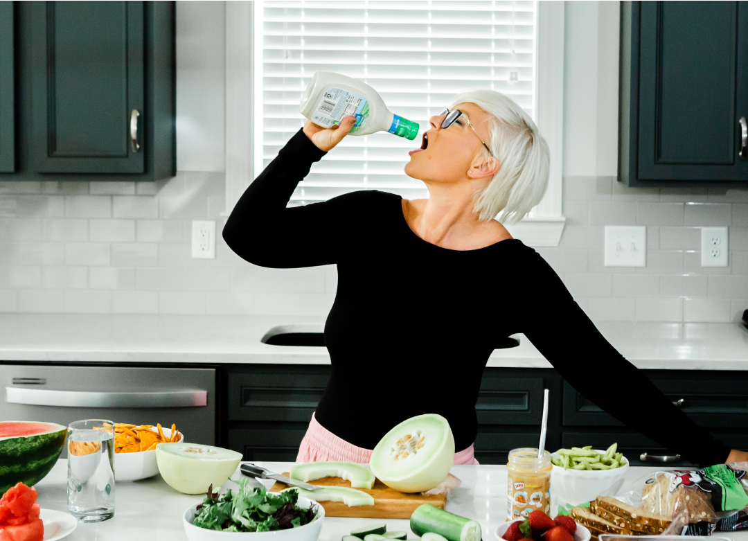 A woman drinking water from a bottle in front of a kitchen full of food.